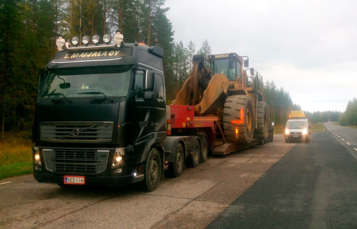 Musta kuorma-auto vetää suurta oranssia työmaa-ajoneuvoa tien laidassa, ja toinen ajoneuvo on takana.