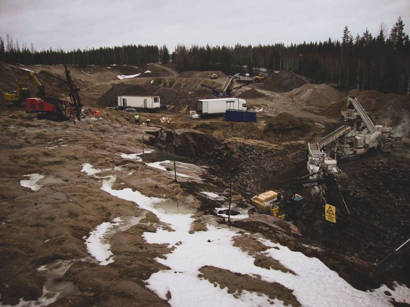 Rakennustyömaa, jossa on kaivinkoneita, hiekkateitä, perävaunuja ja lumilaikkuja metsämaisemassa