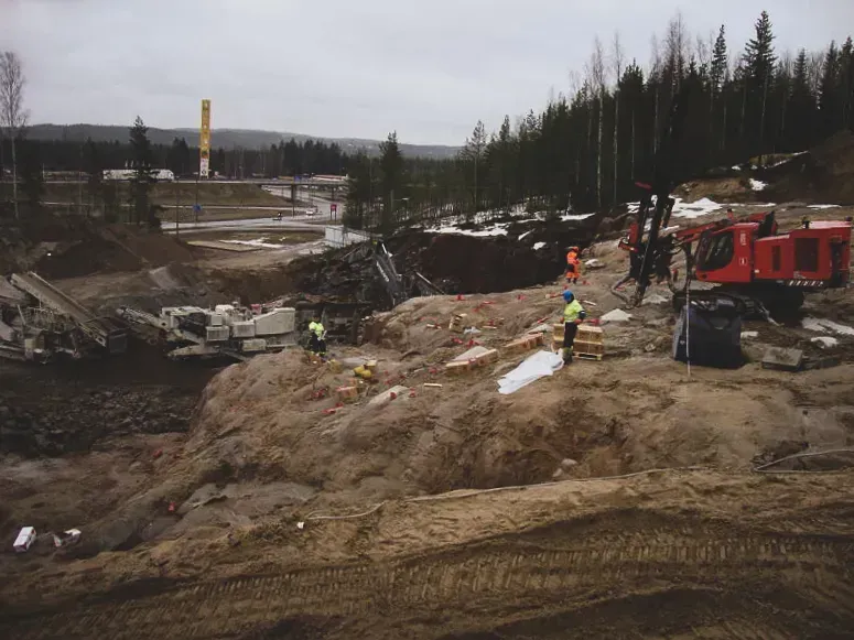 Rakennustyömaa, jossa työntekijöitä heijastinvarusteissa lähellä kaivettua mutaista rinnettä ja punaisia ​​koneita