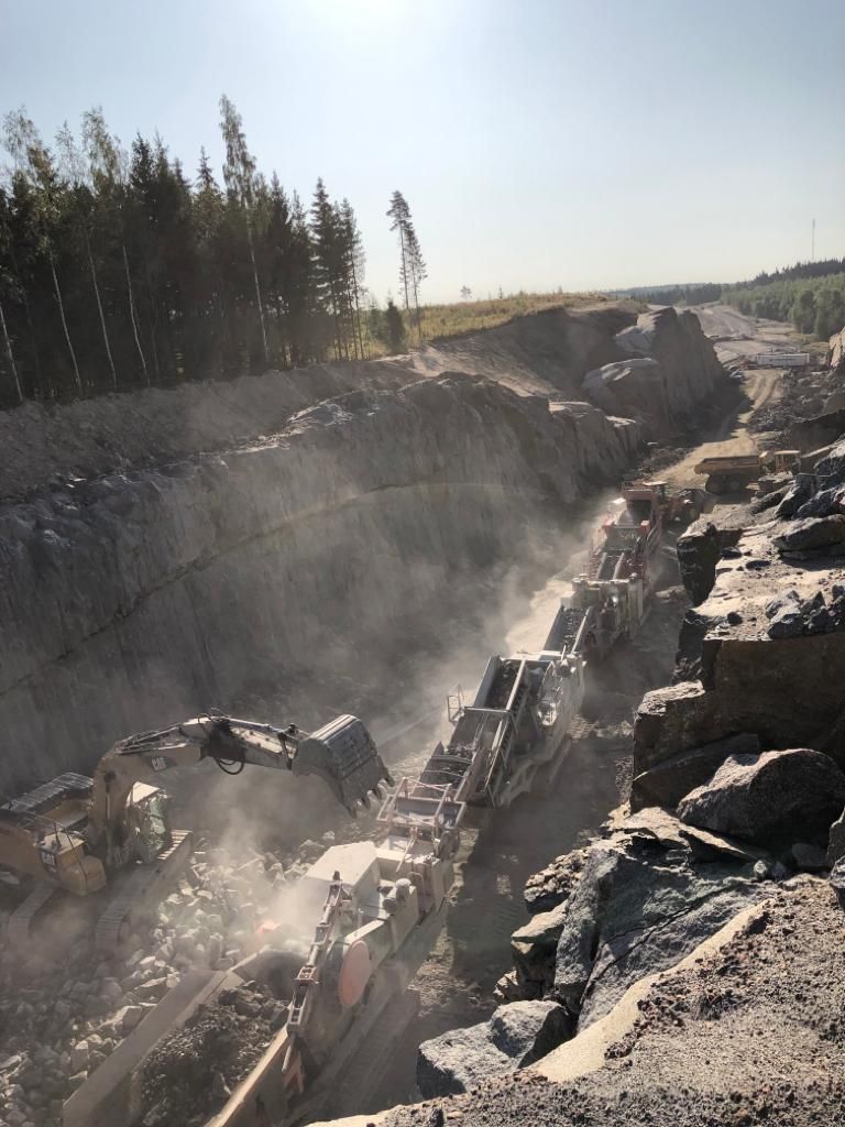 Kivinen louhos, jossa on raskaita koneita ja pölyä syvässä kaivannossa, puut reunustavat horisonttia