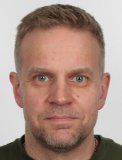 Lähikuva miehestä, jolla on lyhyet vaaleat hiukset ja neutraali ilme, yksiväristä taustaa vasten