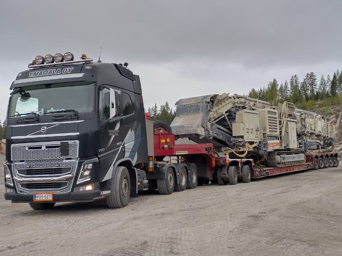 Musta Volvo-puoliperävaunu vetää ylisuuria raskaskalustoa lavetilla soratontilla pilvisen taivaan alla