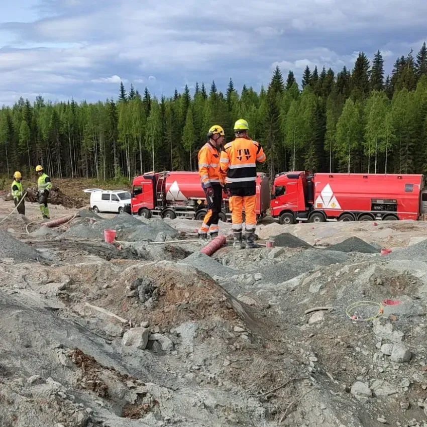 Kaksi oransseihin turvavarusteisiin pukeutunutta työntekijää seisoo punaisten kuorma-autojen vieressä kivisellä rakennustyömaalla, jonka takana on metsää.
