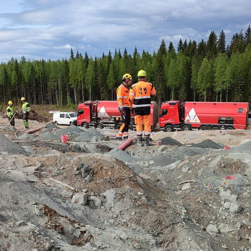 Oranssiin turvavarusteisiin pukeutuneita työntekijöitä punaisen kuorma-auton lähellä kivisellä rakennustyömaalla metsän laidassa