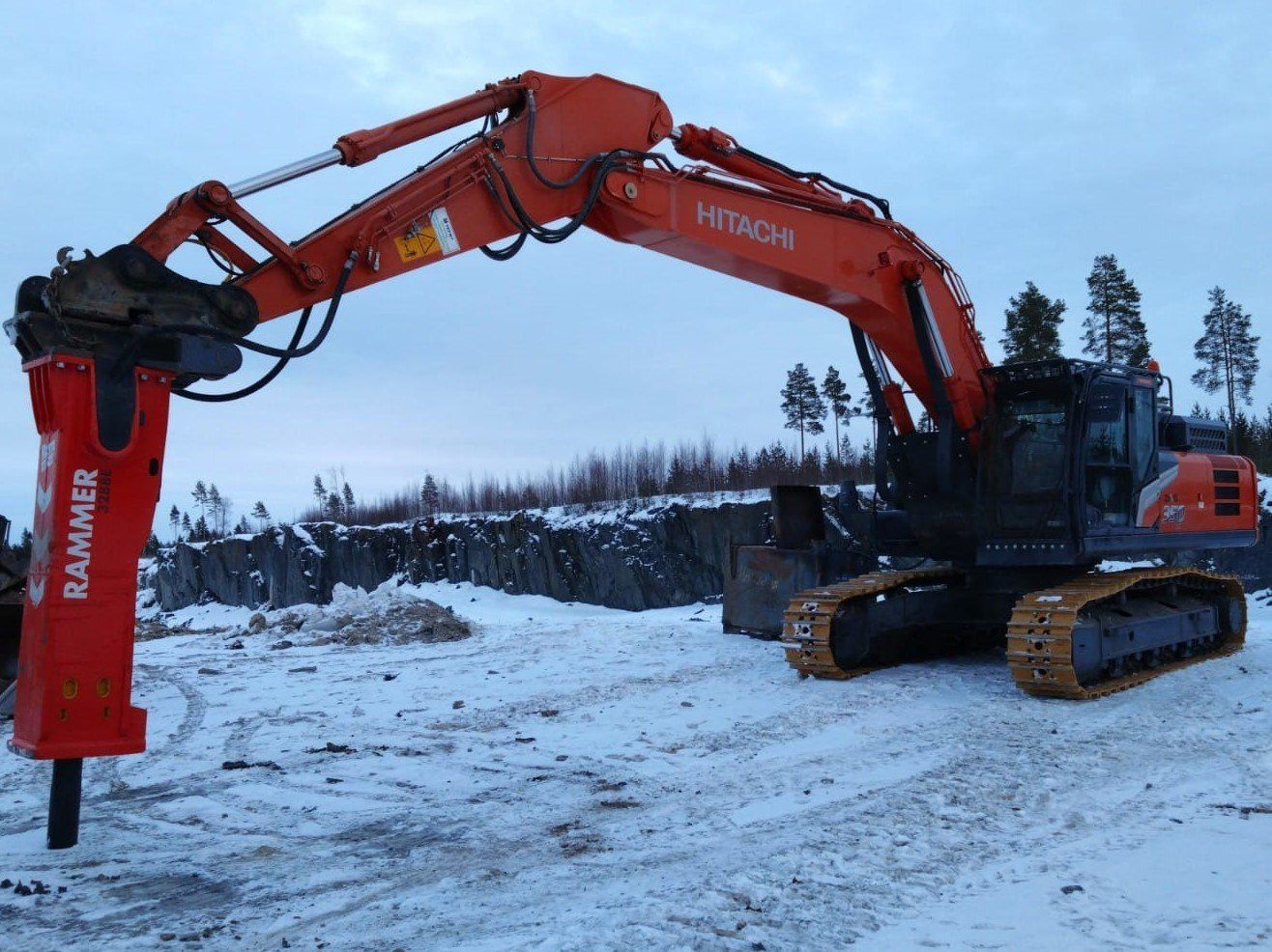 Punainen Hitachi-kaivinkone metsäkoneeseen kiinnitetyllä lisälaitteella lumisella maalla harmaan taivaan alla