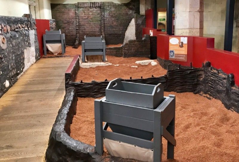 A museum exhibit shows historical grain-processing tools, gray wooden bins, and red walls on a sandy floor.