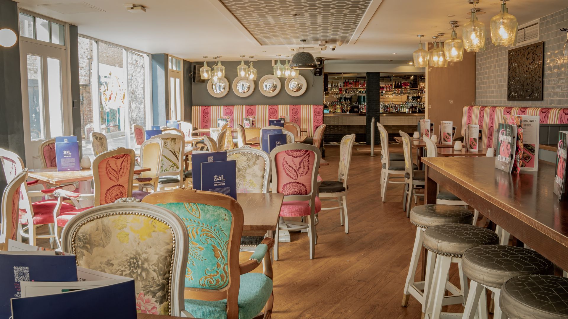 Interior of a colorful restaurant with mismatched chairs, wooden tables, a bar, and large windows.