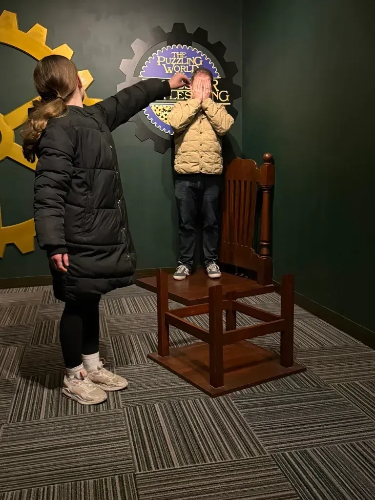 Girl pointing at boy standing on a chair in front of a wall decoration. Both are in a room.