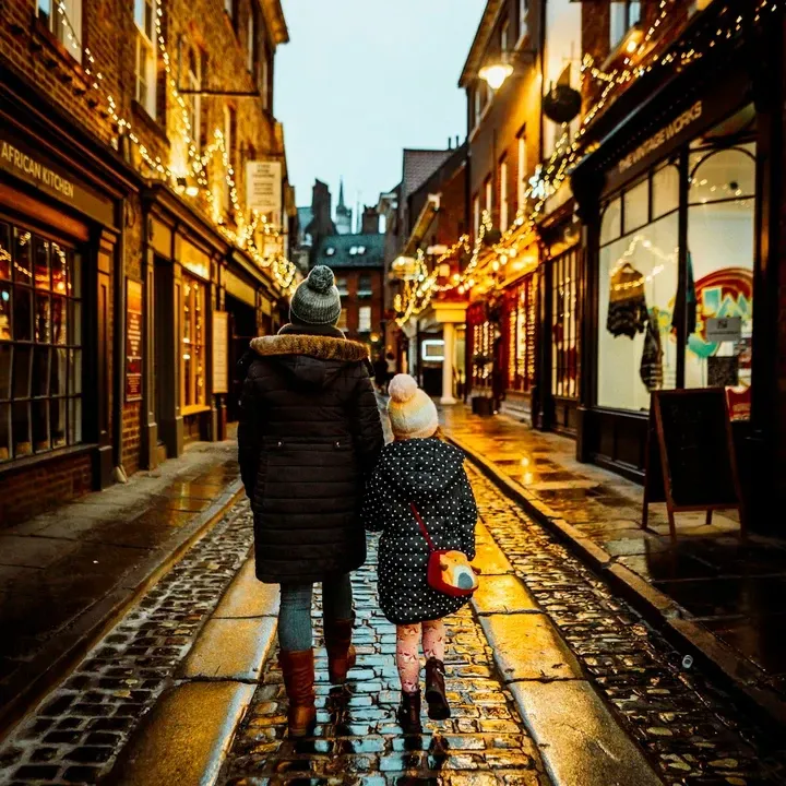 Two people walking down a narrow street lined with shops, illuminated by string lights.