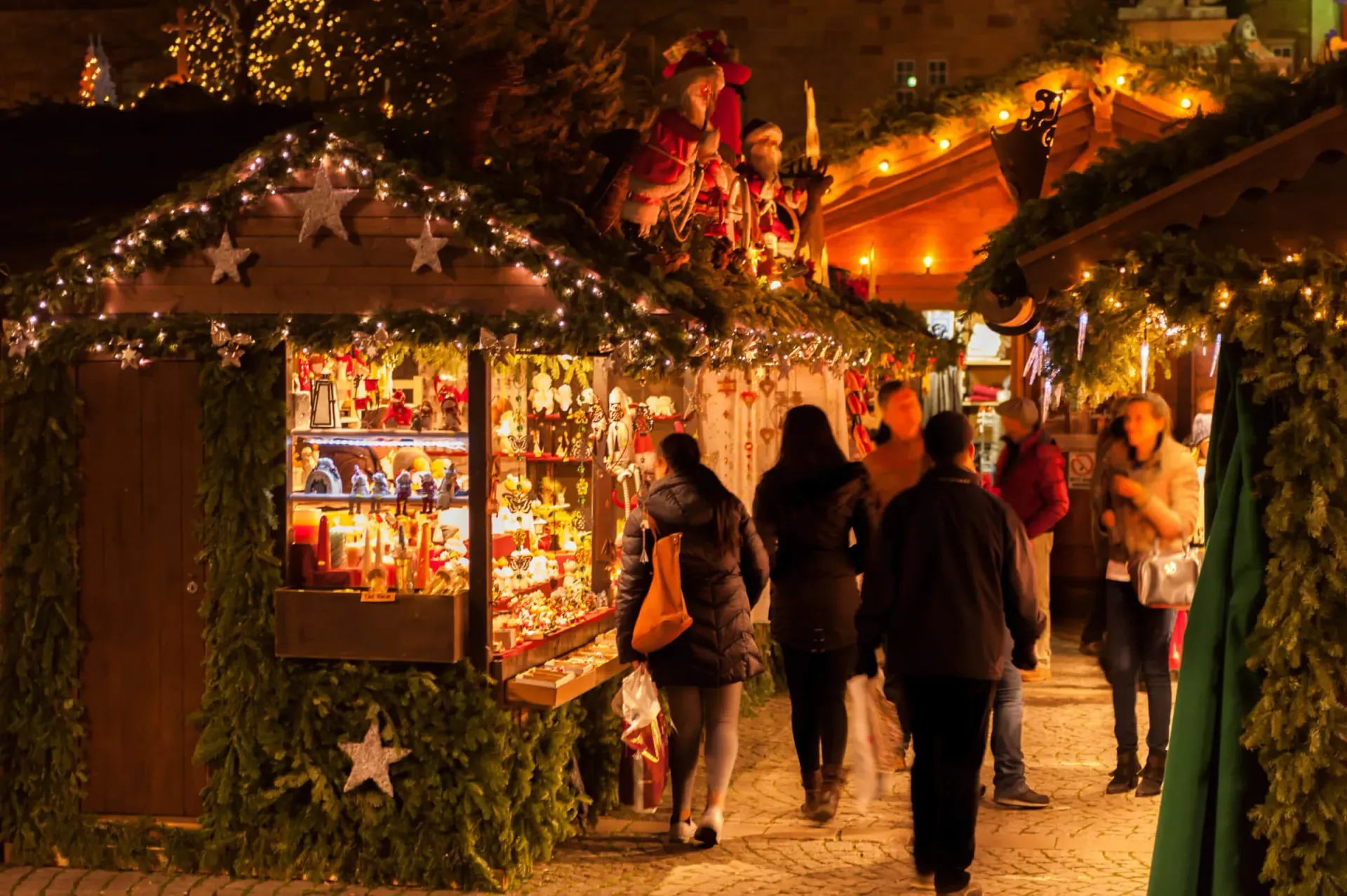 Christmas market at night, people strolling past decorated wooden stalls, illuminated with lights.