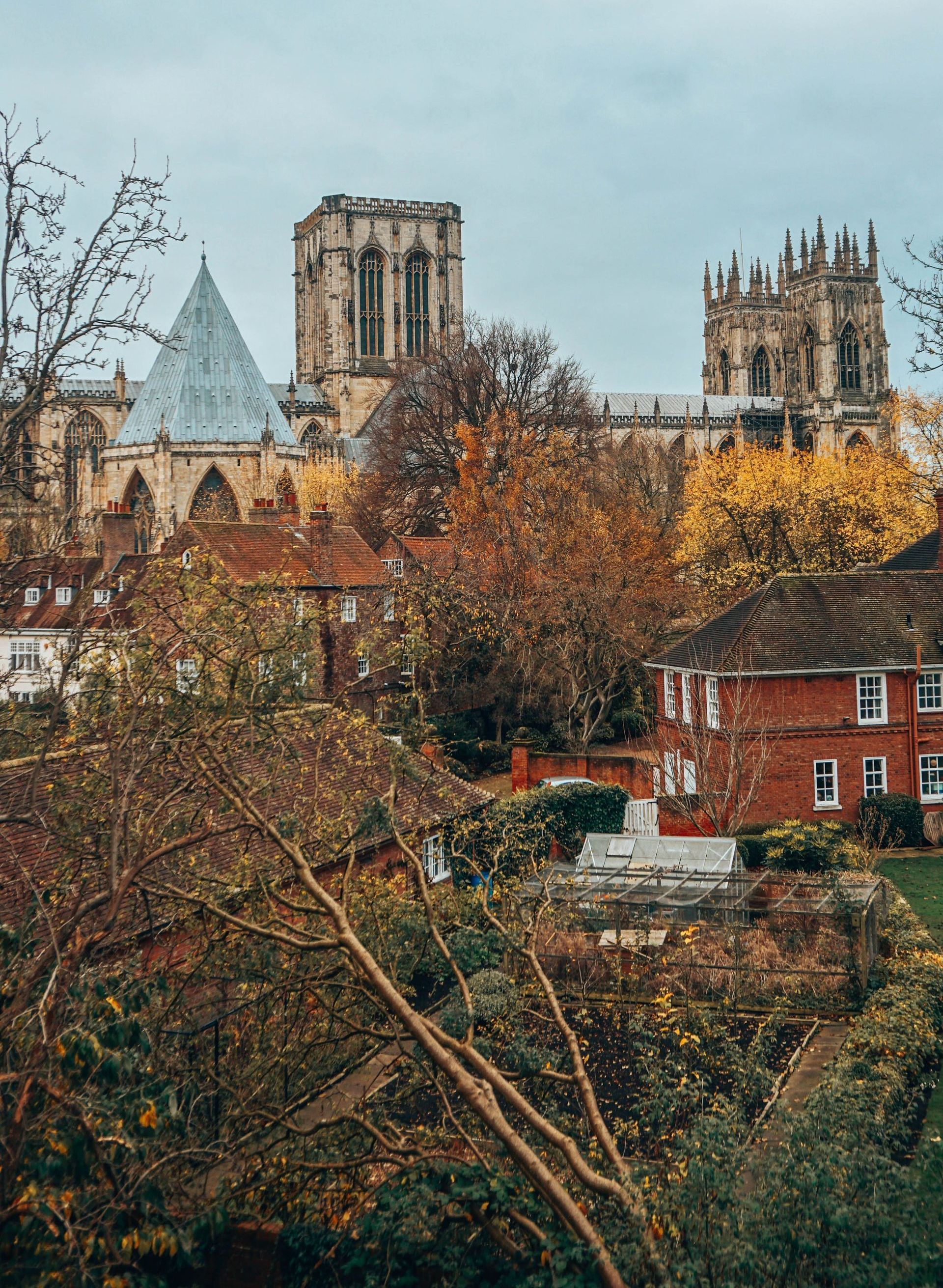 York Minster cathedral rises above autumn trees and red-brick buildings in York, England, under a cloudy sky.