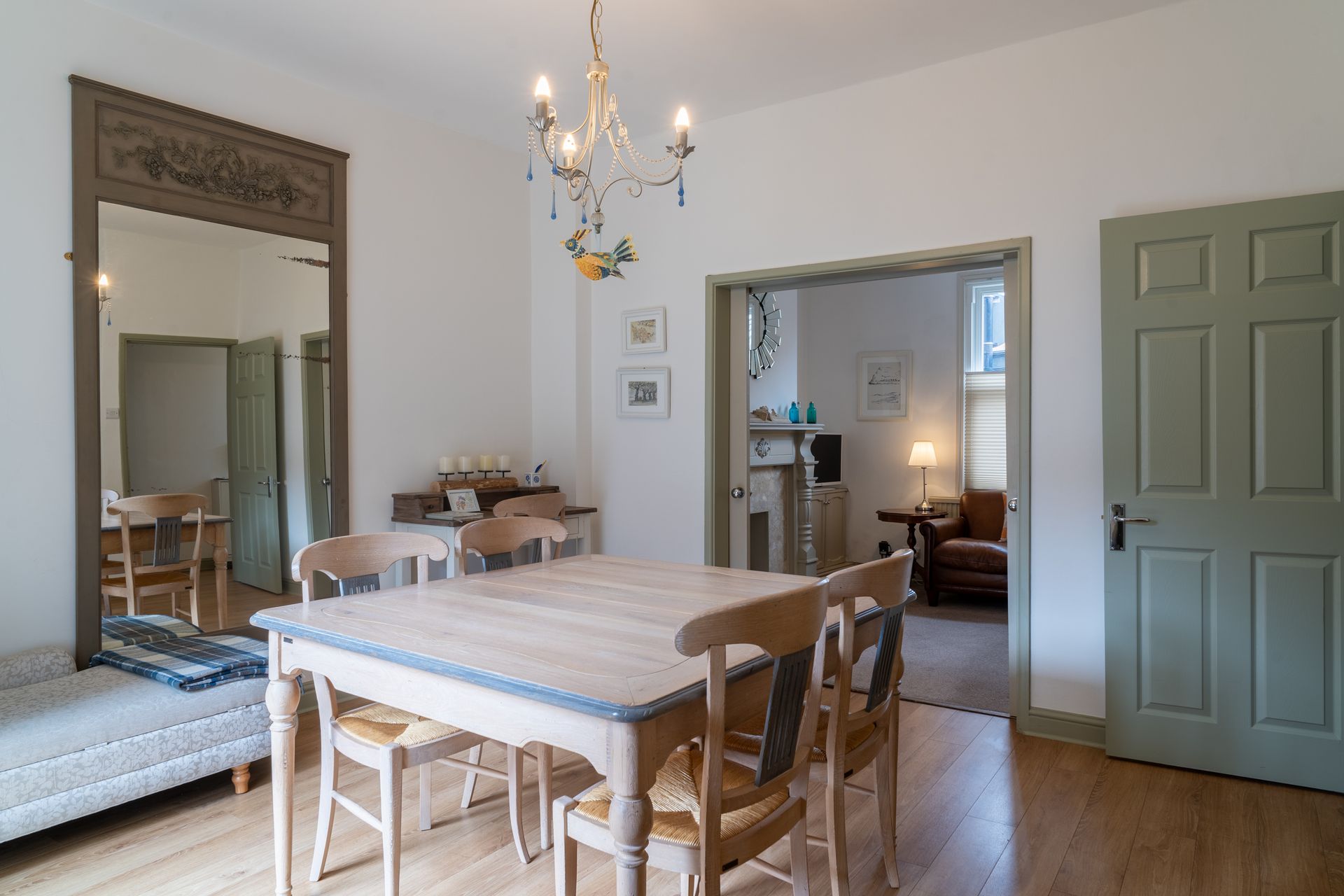 Dining room with a table, chairs, mirror, and a view into a living room. Light-colored walls and floors.