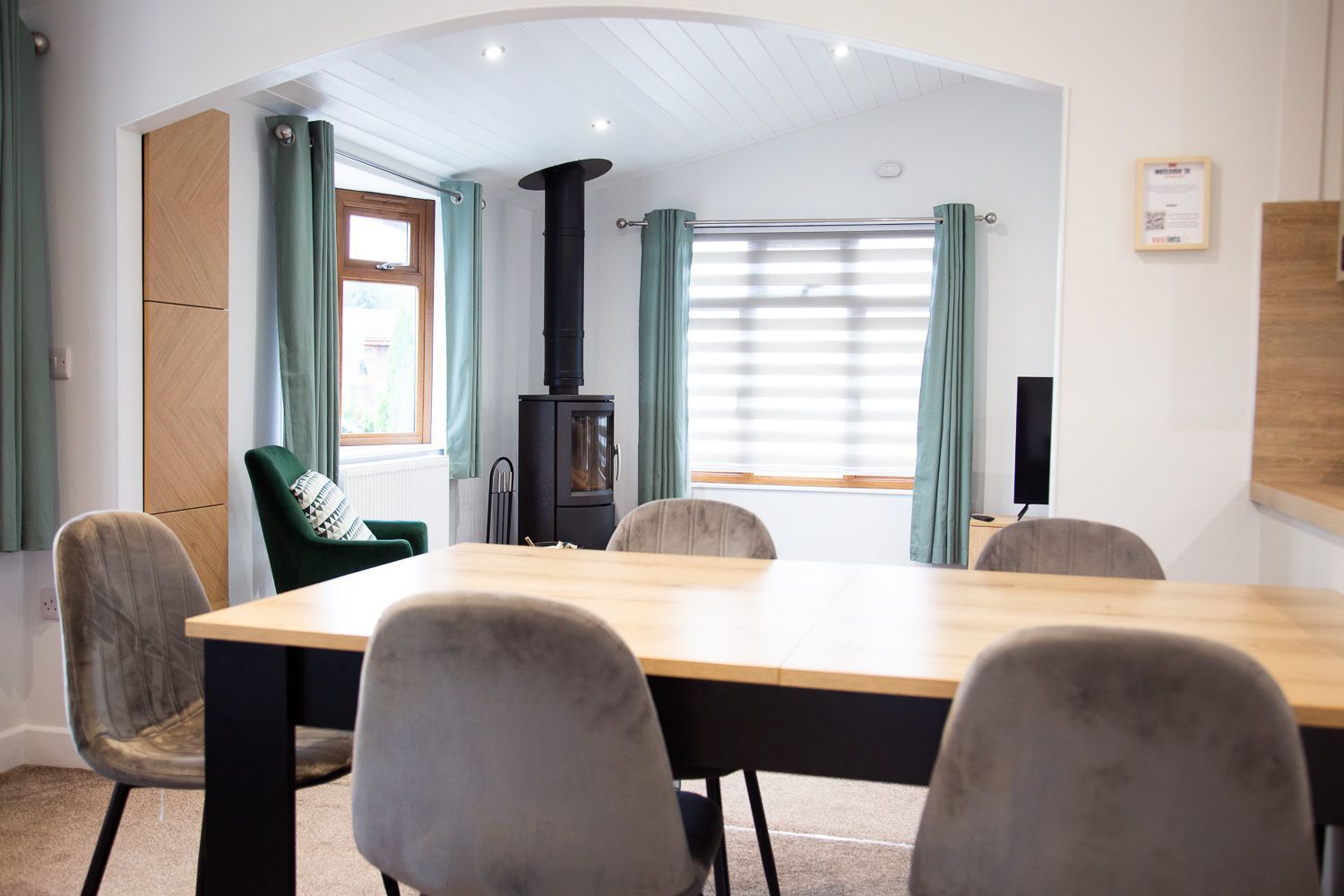 Dining area with wooden table, grey chairs, and fireplace in a cozy, light-filled cabin setting.