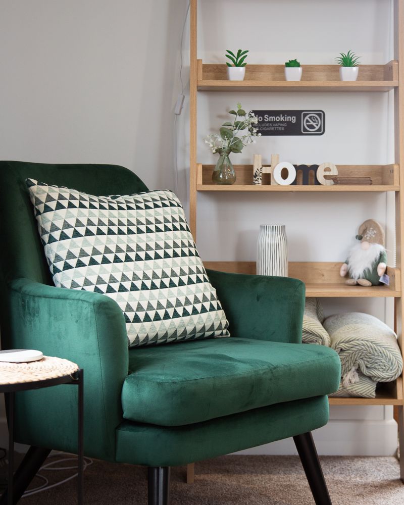 Green velvet armchair with patterned pillow, next to a wooden bookshelf with decor.
