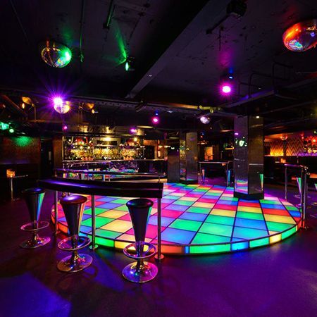 A colorful nightclub dance floor with a bar and disco balls overhead.