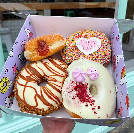Four decorated donuts in a box, including one with sprinkles and a heart-shaped label, held near a window.