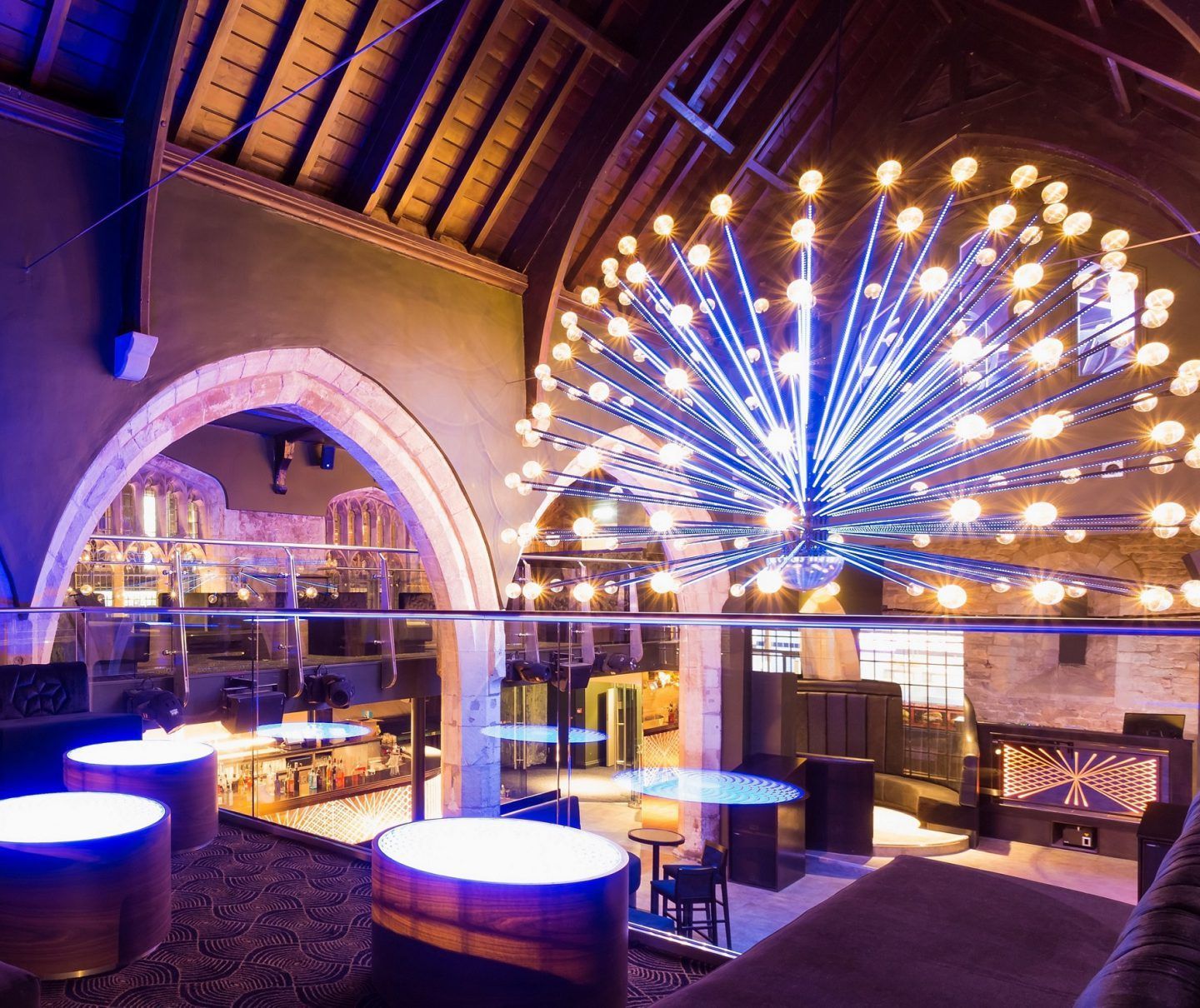 Interior of a lounge, arched ceiling, large light fixture, round tables with blue lighting, fireplace, and seating.
