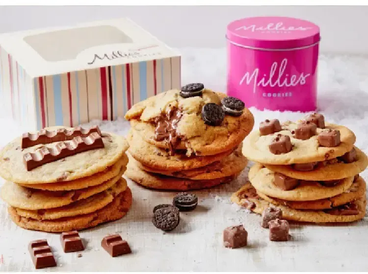 Stacks of cookies with toppings like Kinder, Oreo, and chocolate pieces, with a pink tin and box.