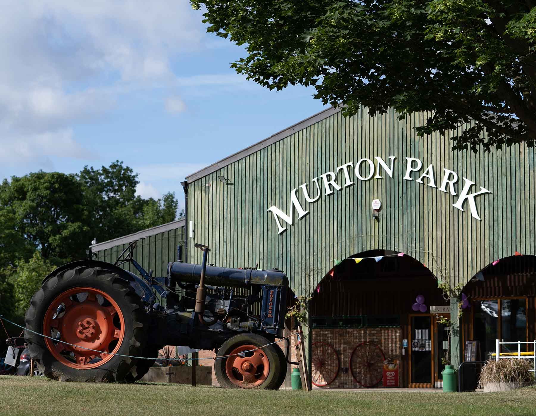 Tractor parked in front of Murton Park entrance, a green barn with white lettering. Bright day, blue sky.