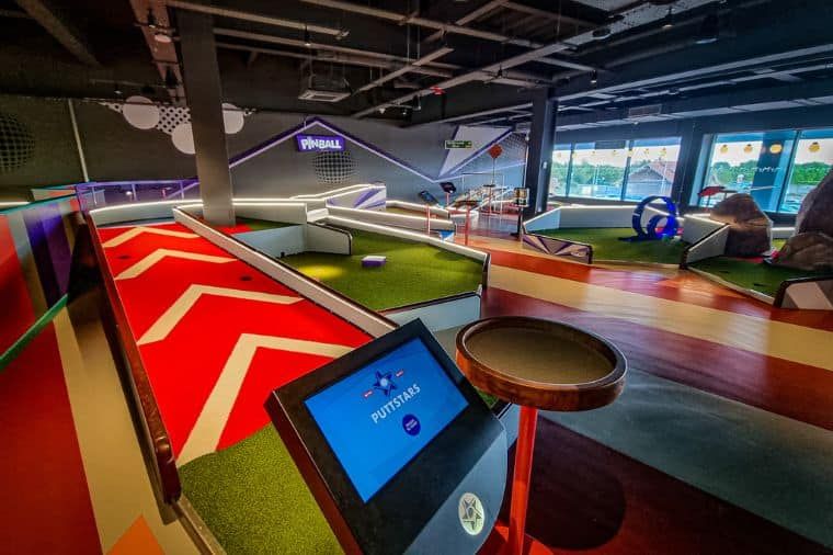 Indoor mini golf course with red and white striped walls, a screen displaying game info, and colorful obstacles.