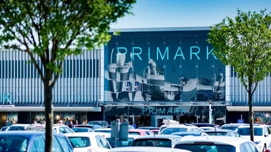 Primark store exterior with logo above entrance, cars in parking lot, and trees.