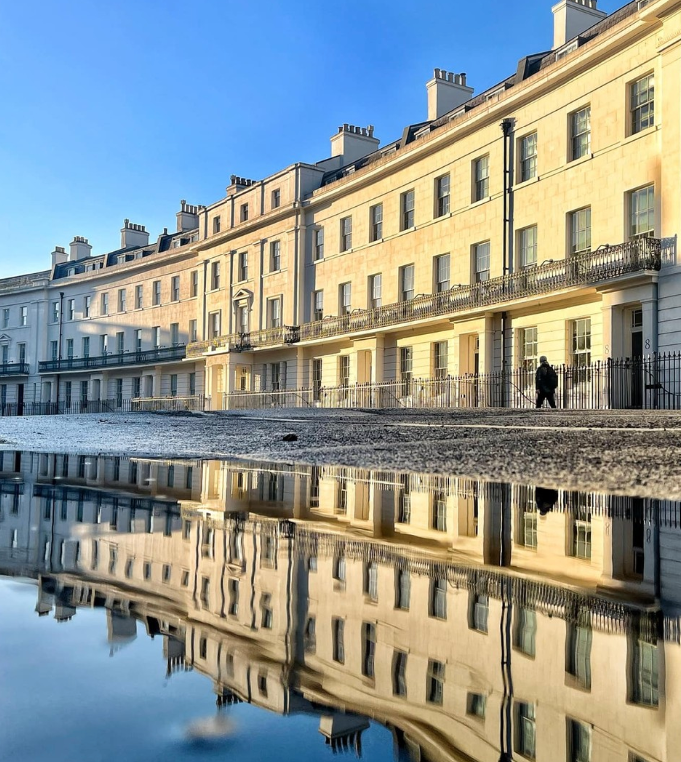 Elegant cream-colored terraced buildings reflect in a puddle on a sunny day; a person walks along the sidewalk.