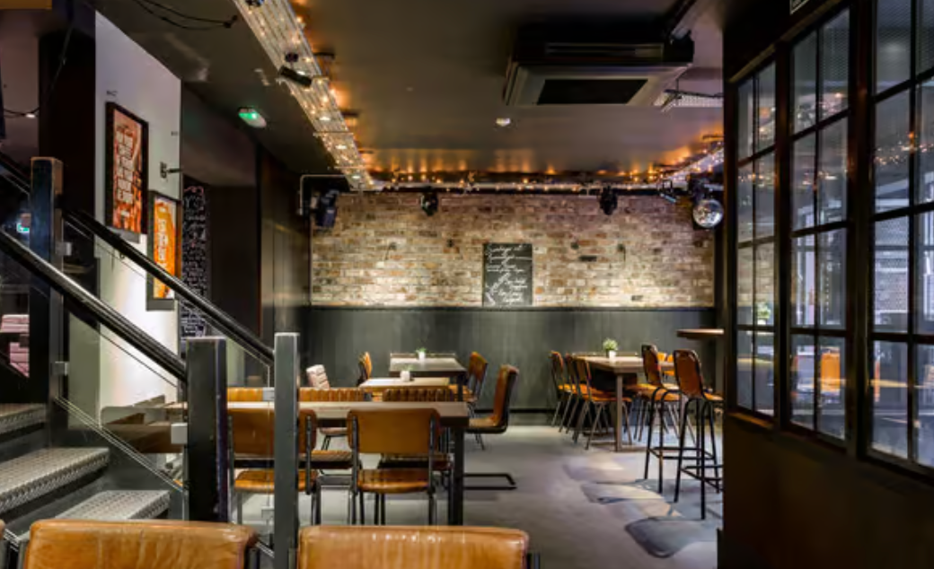 Interior of a pub with tables, chairs, and a brick wall backdrop, with a stairway to the left.