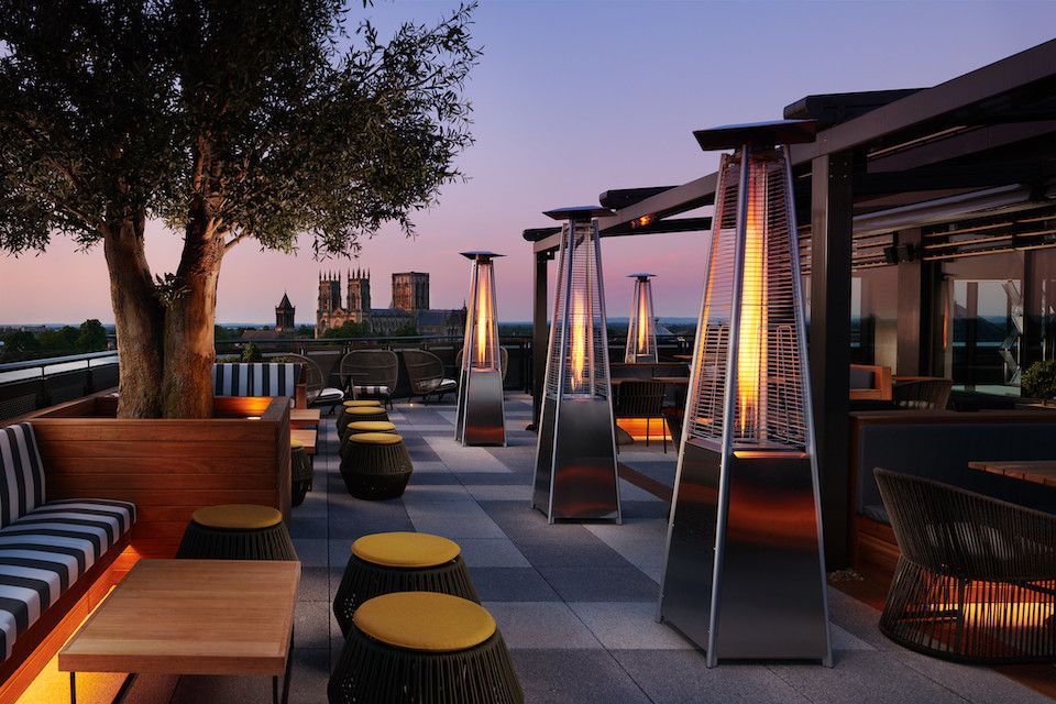 Rooftop patio with heaters, seating, and city view at dusk; tree and pergola.