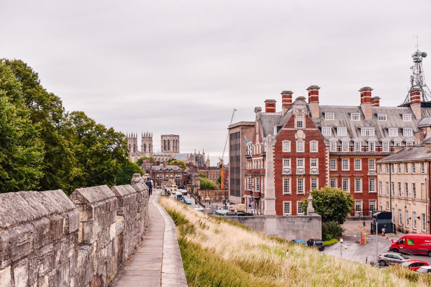Stone city walls with path, buildings, cars, and a distant cathedral in York, England.
