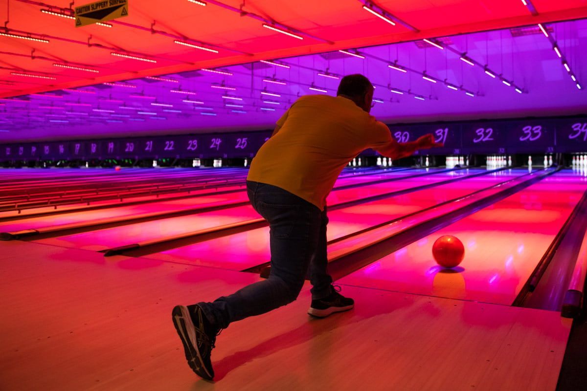 Man bowling in a dimly lit bowling alley with colorful lights. He is wearing a yellow shirt and jeans.