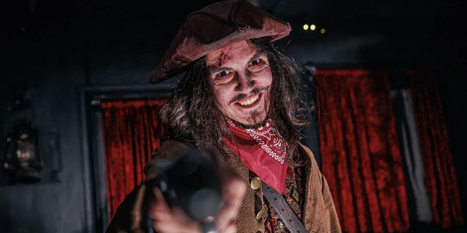 A deranged-looking pirate smiles, holding a pistol. Interior of a ship with red curtains in the background.