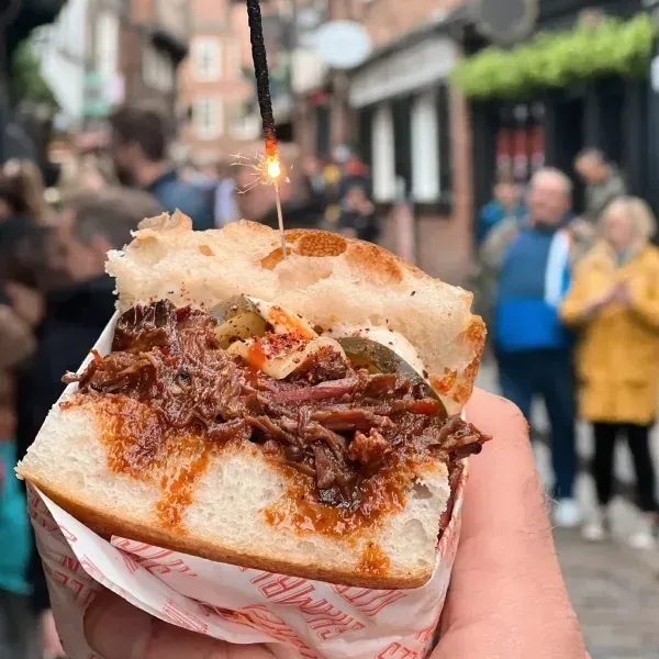 Hand holding a sandwich with meat and pickles, a sparkler burning above, in a crowded outdoor area.