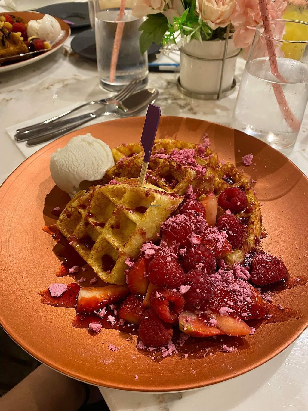 Waffles with berries, ice cream, and pink sprinkles on a copper-colored plate in a restaurant setting.