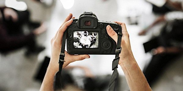 Hands holding a black camera, taking a photo of blurred figures in a light-filled space.