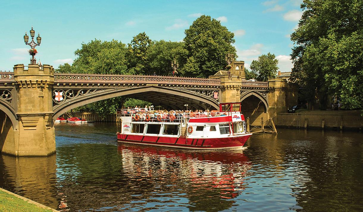 A red tour boat sails under a stone bridge on a river, York, England.