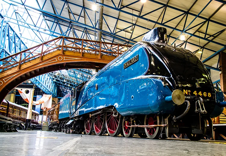 A blue train is parked under a bridge in a building.