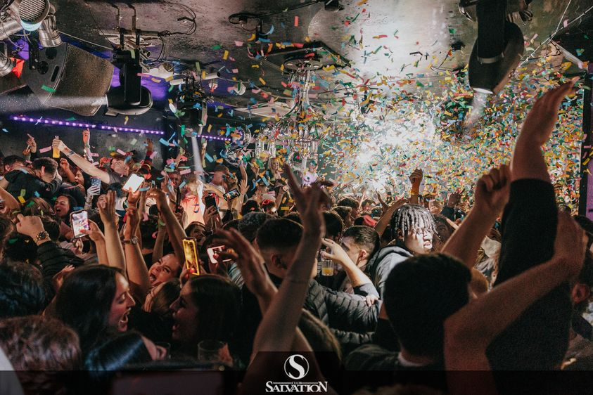 Party crowd with confetti falling, arms raised. Nightclub interior, logo visible.