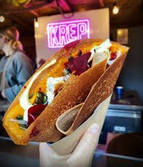 Hand holding a savory crepe filled with colorful ingredients, in front of a 