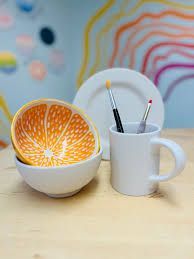 White ceramic dishware and mug with paintbrushes, orange-slice bowl, and colorful wall art on a wooden table.