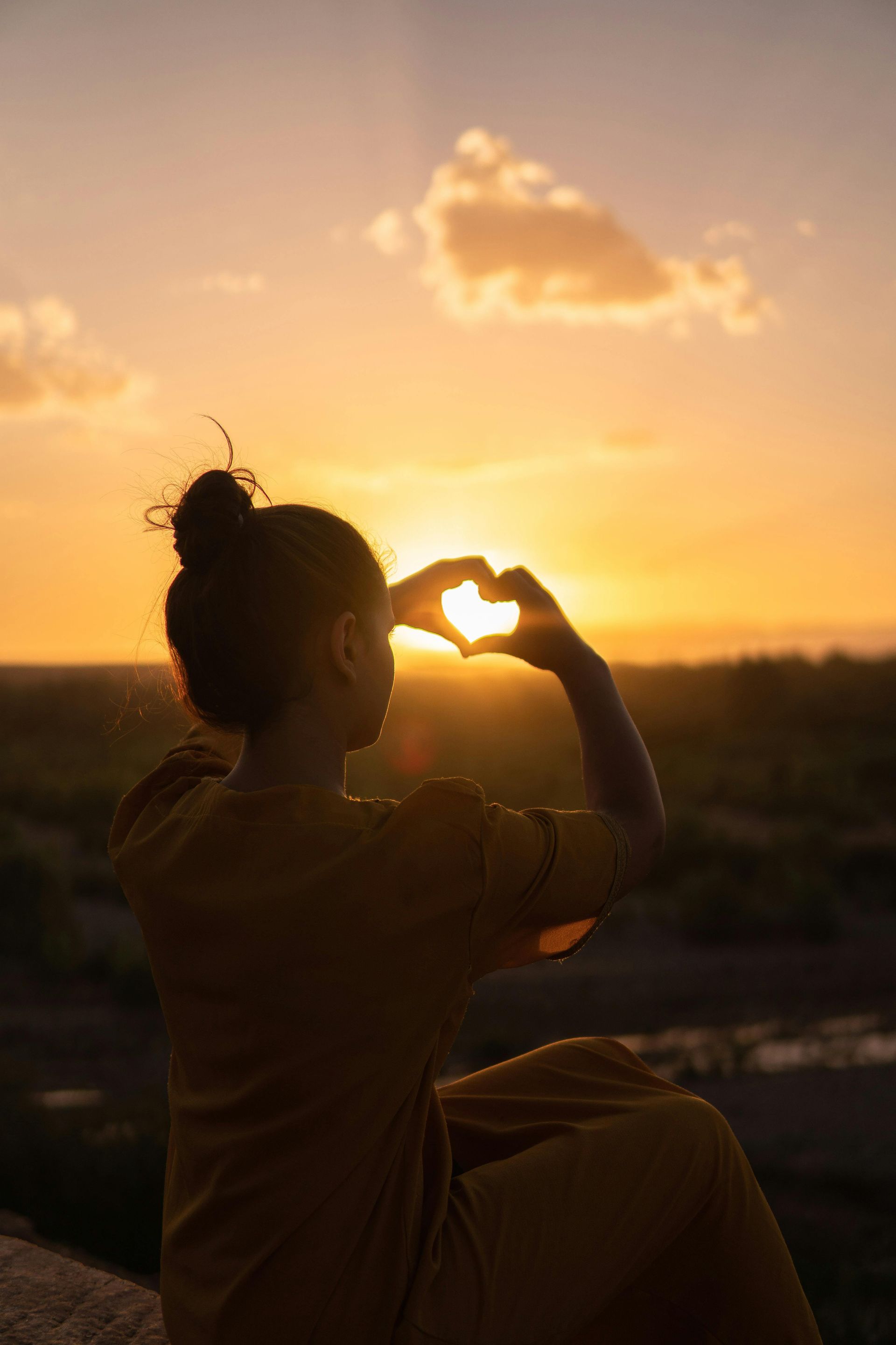 Woman forms a heart with her hands framing the setting sun.