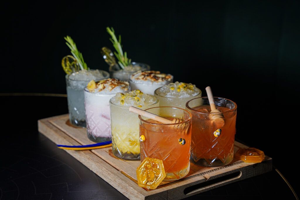Cocktail flight with various drinks on a wooden tray, garnished with citrus and herbs. Dark background.