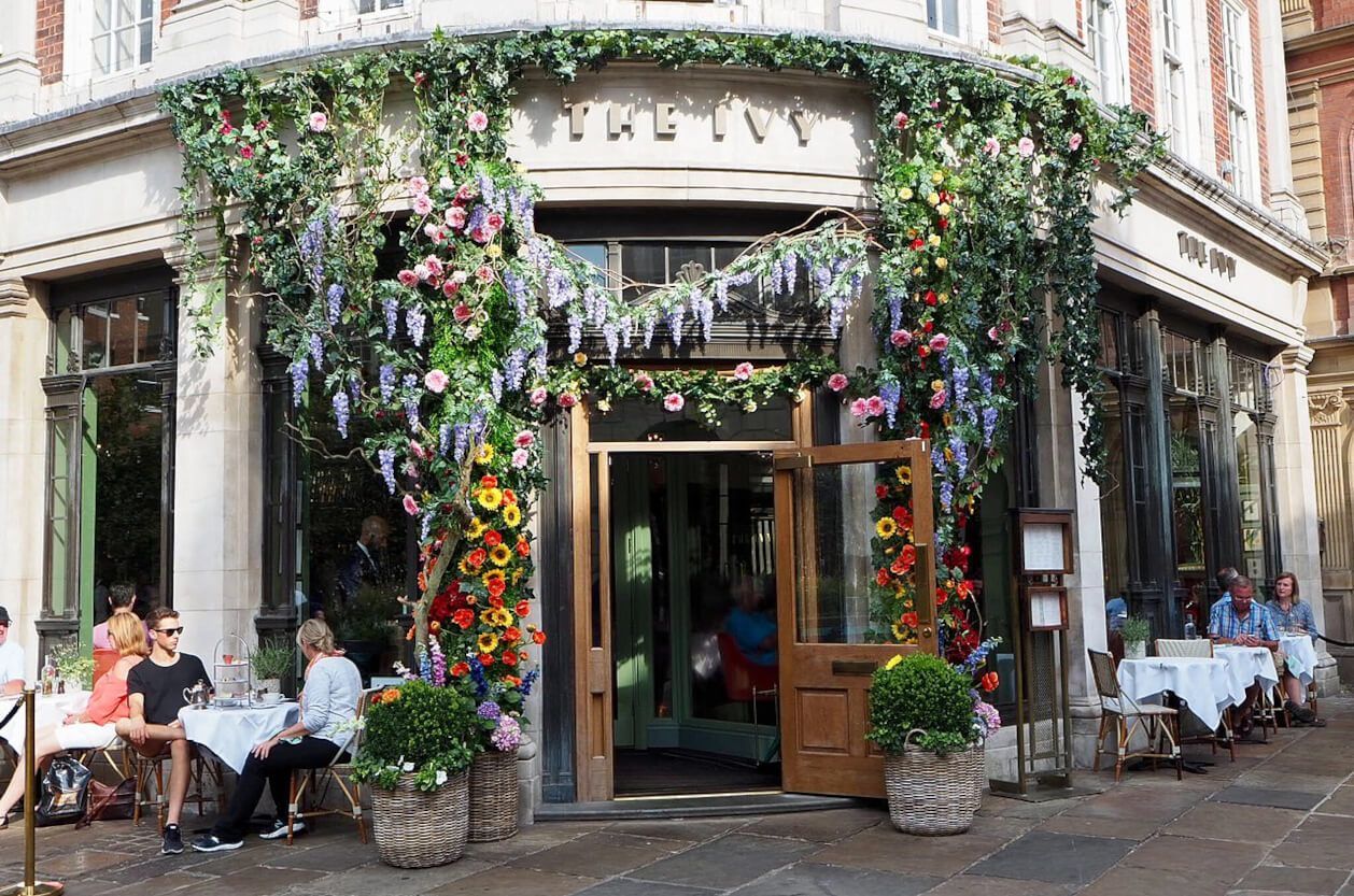 Restaurant entrance, The Ivy, covered in flowers. People seated at tables outside.