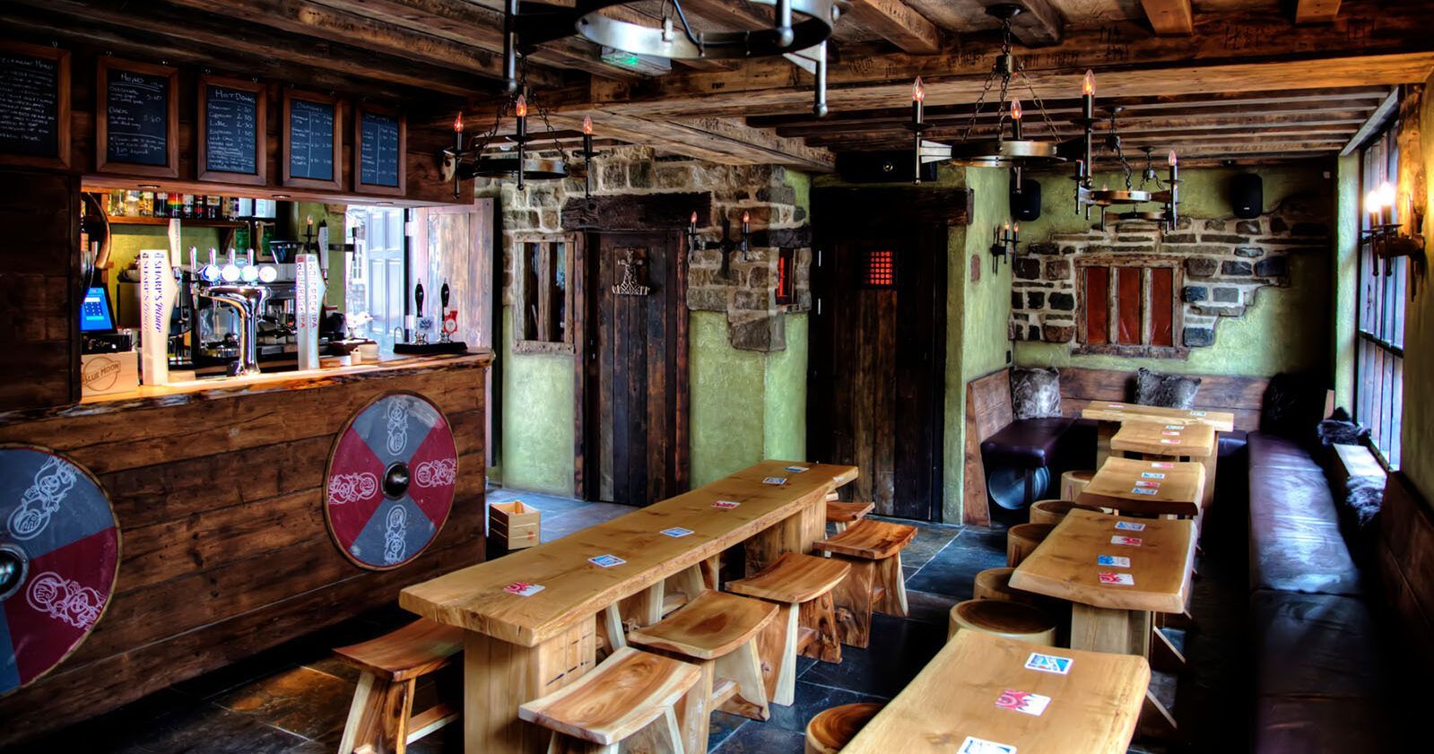 Rustic pub interior: wooden tables, bar, stools, dim lighting, shields on bar.