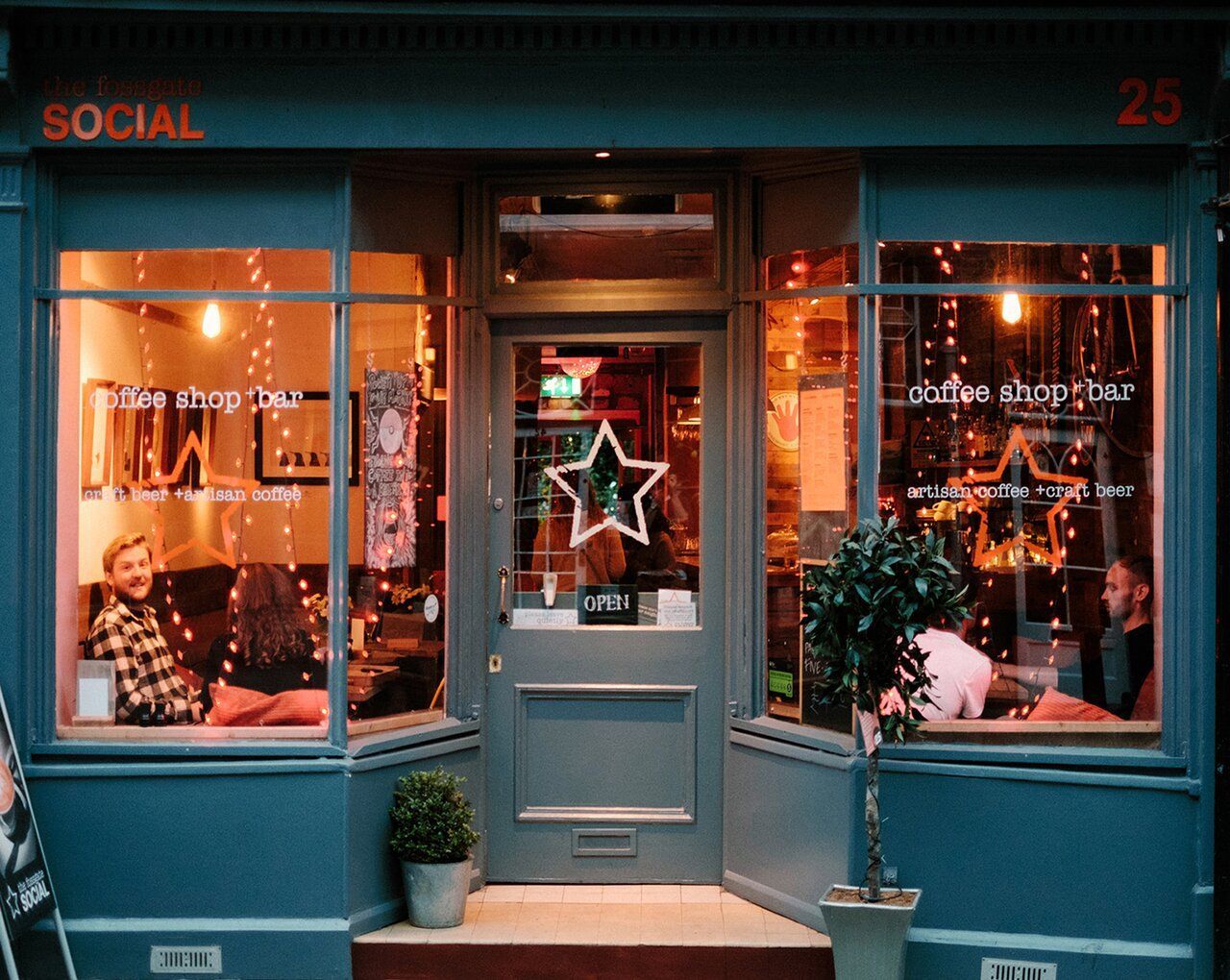 Cafe shop exterior with teal paint, Christmas lights in windows, open door with star. People inside.