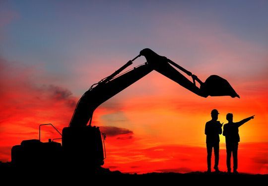 Dos hombres están parados frente a una excavadora al atardecer.