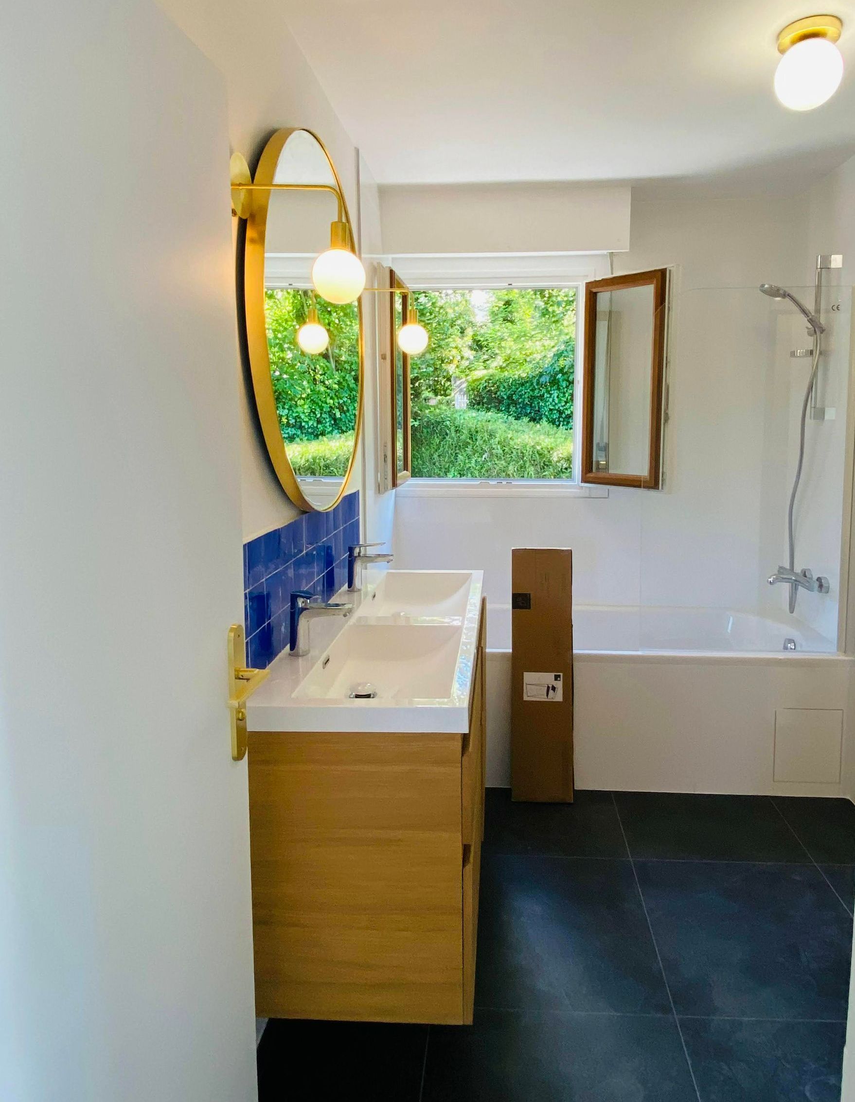 Salle de bains avec meuble-lavabo en bois, miroir doré et fenêtre ouverte donnant sur une végétation luxuriante.
