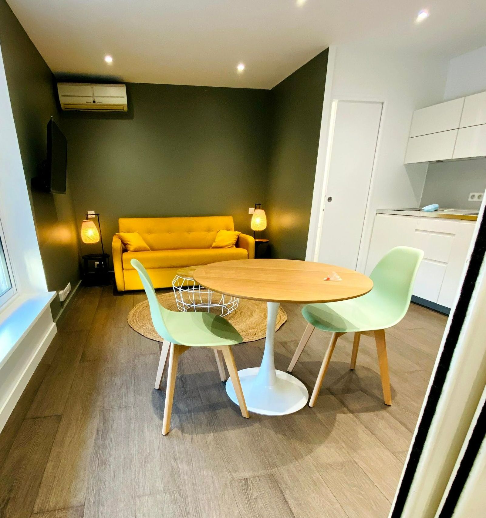 Intérieur d'un petit appartement avec des murs vert olive, un canapé jaune, une table ronde et des chaises vert clair.