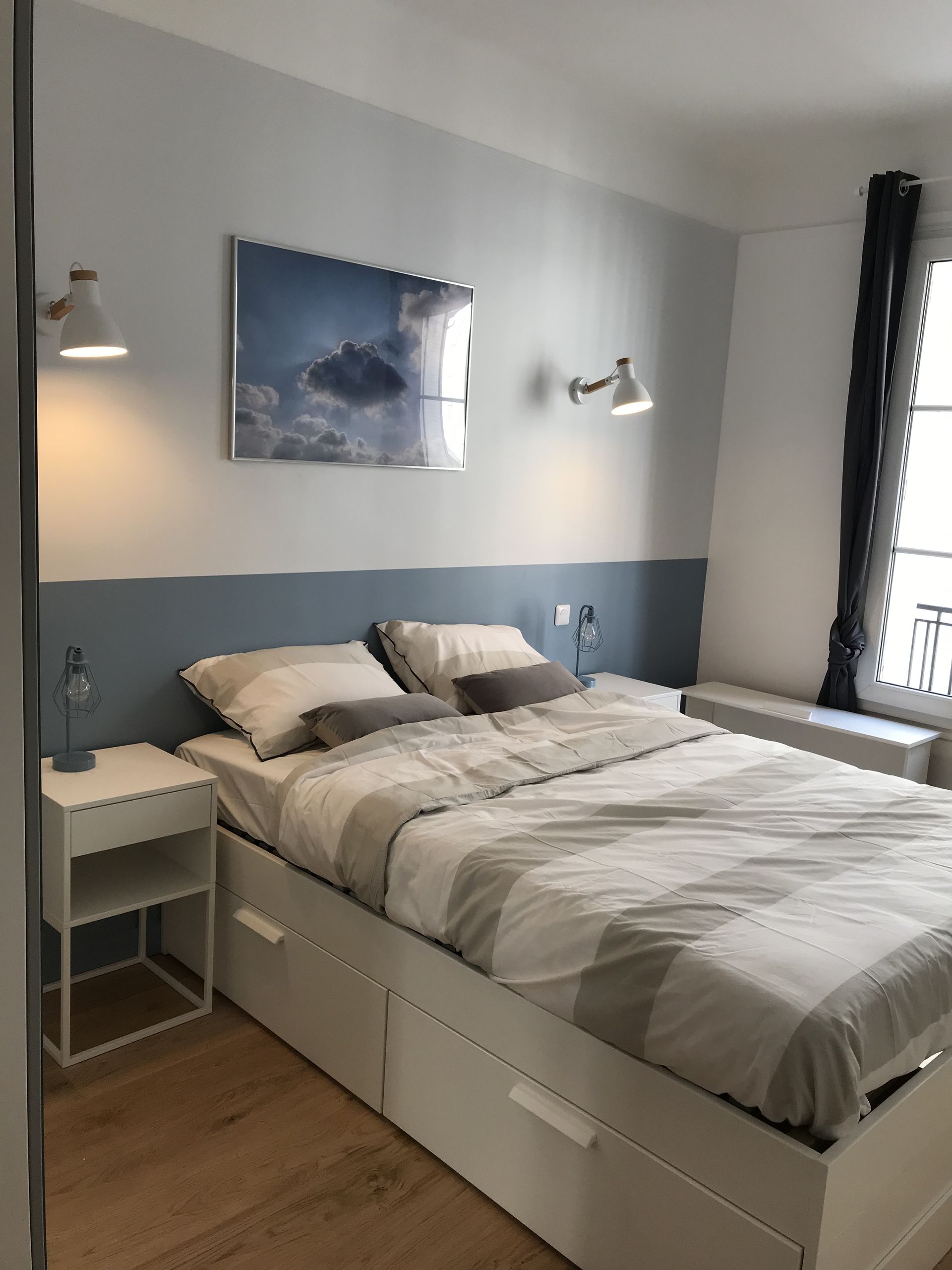 Chambre avec lit blanc, mur bleu et blanc, table de chevet, tableau et fenêtre.