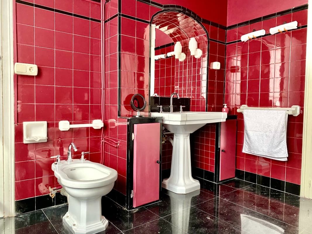 Baño público con azulejos rojos, inodoro, lavabo con pedestal, espejo y toallero.