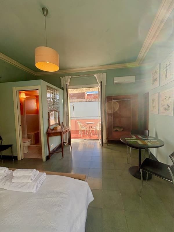 Habitación luminosa con cama, sillas, puerta de balcón abierta y paredes de un suave color verde.